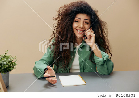 Happy smiling tanned curly Latin lady talking with friend in call with phone sit at the table isolated over pastel beige background. Copy space Mockup Banner. Concept cellular communication Happy smiling tanned curly Latin lady talking with friend in call with phone sit at the table isolated over pastel beige background. Copy space Mockup Banner. Concept cellular communication 90108301
