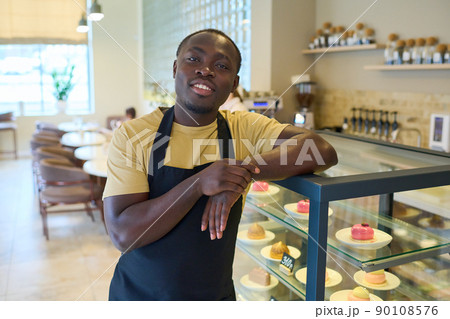 Portrait of African young owner in black uniform leaning on counter with sweet food and smiling at camera during his work in coffee shop Portrait of African young owner in black uniform leaning on counter with sweet food and smiling at camera during his work in coffee shop 90108576