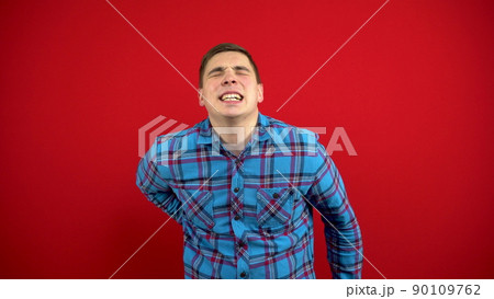 A young man has a backache and is holding it in his hands. Shooting on a red background. 90109762