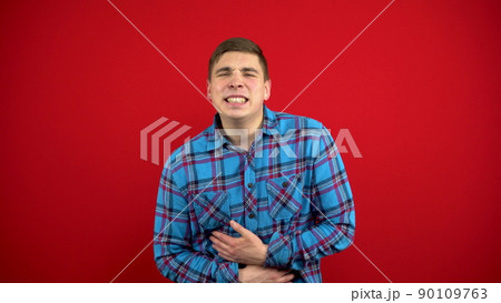 A young man has a stomach ache and he holds it in his hands. Shooting on a red background. 90109763