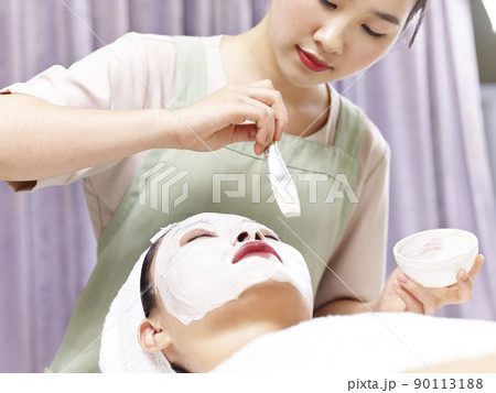 asian beautician applying white facial mask with a brush on face of a young woman asian beautician applying white facial mask with a brush on face of a young woman 90113188