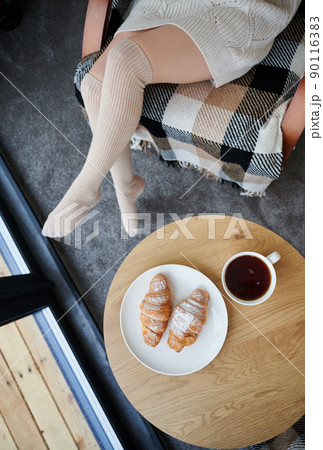 Top view of young woman's legs near window inside contemporary barn house. Place with croissants and cup of tea on the table. 90116383