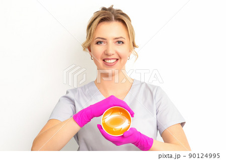 Portrait of a female caucasian beautician holding a jar of sugar paste for sugaring wearing pink gloves on white background 90124995