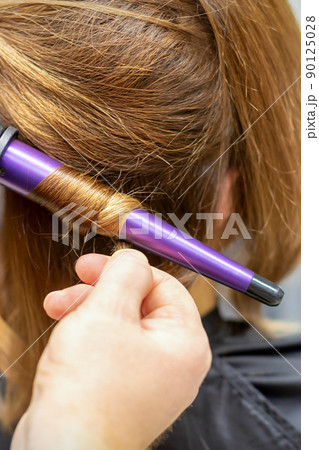 The hairstylist makes curls hairstyle of long brown hair with the curling iron in hairdresser salon, close up 90125028