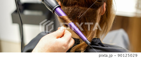 The hairstylist makes curls hairstyle of long brown hair with the curling iron in hairdresser salon, close up 90125029