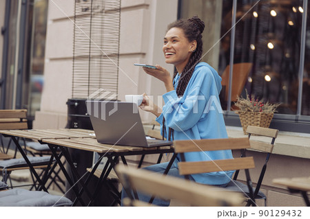 Pretty cheerful young woman with laptop sending voice message and having coffee at cafe terrace. 90129432