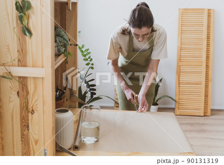 Woman seller in a green aprone cuts craft paper with scissors. Flower and gift shop Woman seller in a green aprone cuts craft paper with scissors. Flower and gift shop 90131319