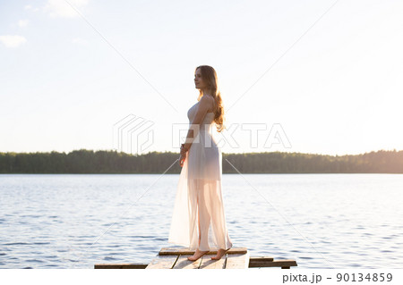 Beautiful Russian girl dressed in a white dress, walking along a wooden pier on the bank of a river or a lake Beautiful Russian girl dressed in a white dress, walking along a wooden pier on the bank of a river or a lake 90134859