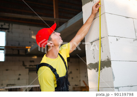 Construction worker at construction site measures the length of window opening and brick wall with tape measure. Cottage are made of porous concrete blocks, work clothes - jumpsuit and baseball cap 90139798