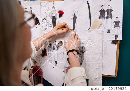 Fashion designer's hands holding pencil and sketching clothes on paper pinned to the wall with drawings and esquises attached. Fashion designer's hands holding pencil and sketching clothes on paper pinned to the wall with drawings and esquises attached. 90139922