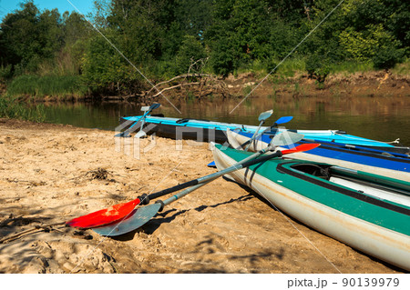 Tourist canoes with paddles stand on the river coast in summer on a water hike. Rafting on inflatable and frame double and triple kayak boats, family trip, extreme adventure in summer Tourist canoes with paddles stand on the river coast in summer on a water hike. Rafting on inflatable and frame double and triple kayak boats, family trip, extreme adventure in summer 90139979