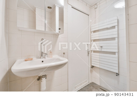 Cupboard with sink and mirror attached to tiled wall near door and drying rack in modern restroom Cupboard with sink and mirror attached to tiled wall near door and drying rack in modern restroom 90143431