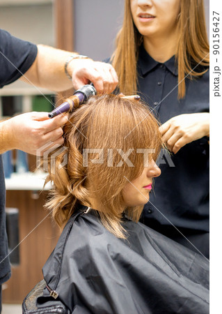 Two hairstylists make curls hairstyle of long brown hair with the curling iron in hairdresser salon 90156427