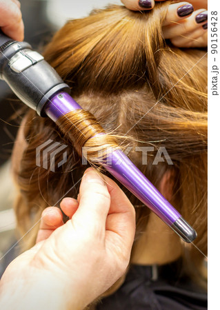 The hairstylist makes curls hairstyle of long brown hair with the curling iron in hairdresser salon, close up 90156428