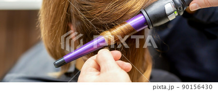 The hairstylist makes curls hairstyle of long brown hair with the curling iron in hairdresser salon, close up 90156430