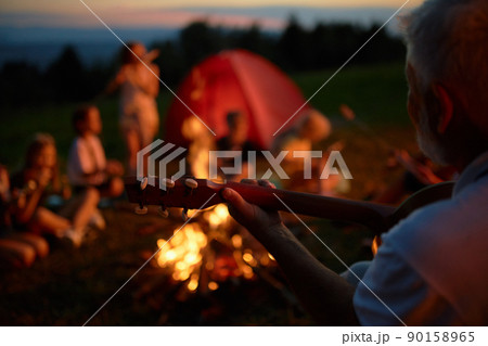 Blurred group of teen friends sitting around campfire, having fun in mountains. Crop view of adult bearded man playing guitar and singing to kids, while camping in evening. Concept of camping. Blurred group of teen friends sitting around campfire, having fun in mountains. Crop view of adult bearded man playing guitar and singing to kids, while camping in evening. Concept of camping. 90158965