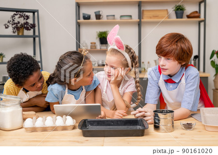 kids wrapped up in reading recipe with help of digital tablet while standing at wooden kitchen table in order to make surprise for Mothers Day kids wrapped up in reading recipe with help of digital tablet while standing at wooden kitchen table in order to make surprise for Mothers Day 90161020