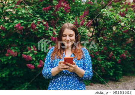 A portrait of a smiling beautiful person texting sms with her phone on nature background. Happy woman is using a smartphone in park outdoors, communications and technology concept 90161632