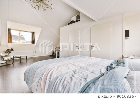 Comfortable bed located against window near wooden armchair and plant and doors in contemporary mansard bedroom 90163267