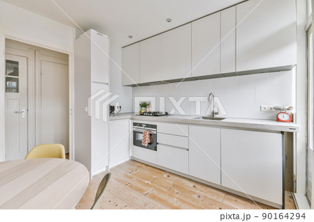 Fragment of interior of light narrow home kitchen with minimalist style white furniture with sink and stove Fragment of interior of light narrow home kitchen with minimalist style white furniture with sink and stove 90164294