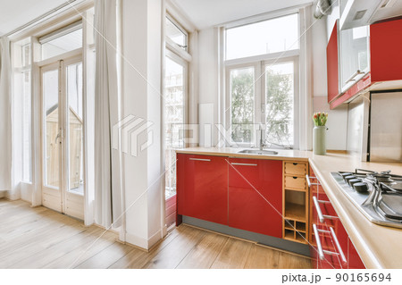 Small corner kitchen with sink under window and red furniture with transparent cupboards with stove near balcony 90165694
