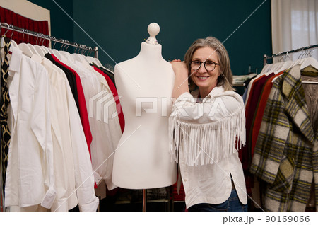 Charming elderly European woman seamstress, tailor, fashion designer posing with a dressmaker's model in her own fashion design atelier Charming elderly European woman seamstress, tailor, fashion designer posing with a dressmaker's model in her own fashion design atelier 90169066