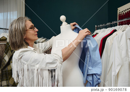Caucasian woman fashion designer, seamstress tries on a blue shirt from new collection on a mannequin in her clothing design and tailoring workshop 90169078