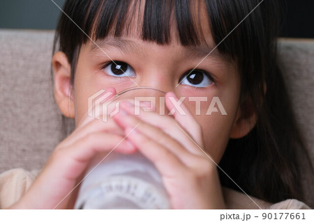 Portrait of a cute Asian little girl holding a glass of milk sitting on the sofa at home. Small girl at home with smiling face, feeling happy enjoying drinking milk and looking at camera. Portrait of a cute Asian little girl holding a glass of milk sitting on the sofa at home. Small girl at home with smiling face, feeling happy enjoying drinking milk and looking at camera. 90177661
