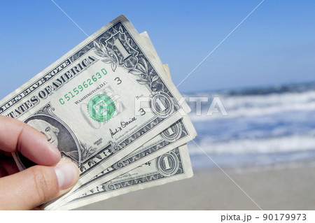 Person holding three paper dollar bills on background of sea and sea waves with white foam, sandy beach and clear blue sky on sunny summer day. Concept of money, expenses on vacation, travel, tourism 90179973