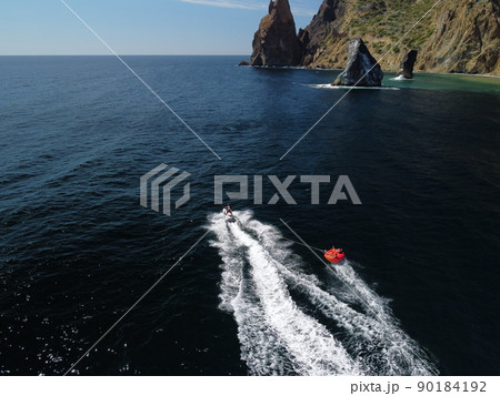 Happy people swim on air mattress behind a high speed water bike. Tourists ride on inflatable watercraft boat. Sea attraction. Water scooter skating people on large Inflatable raft. Aerial copter view 90184192