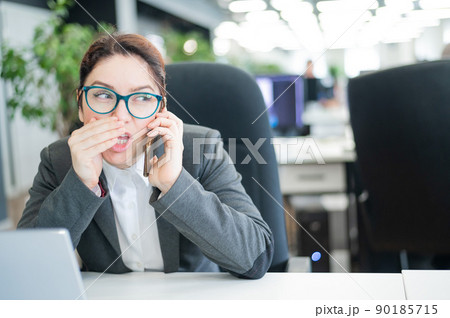 A woman sits in an office at her desk and gossips on the phone. Corporate ethics. Female employee in a suit tells secrets on a smartphone at work. A woman sits in an office at her desk and gossips on the phone. Corporate ethics. Female employee in a suit tells secrets on a smartphone at work. 90185715