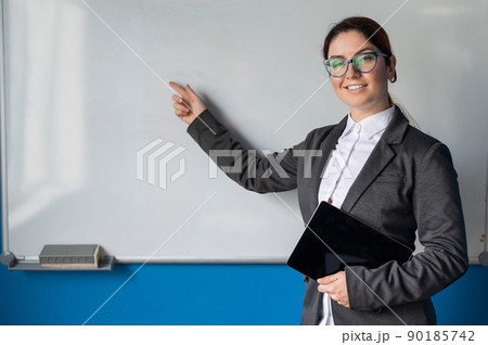 Beautiful woman in a suit with a digital tablet in her hands. A smiling female business coach with glasses stands in a conference room and points a finger at a white board. Workshop at the office. 90185742