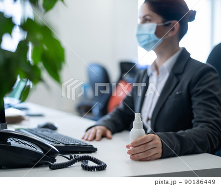 Female office manager in a mask at the desk using an antiseptic to disinfect hands. A woman in a suit processes the hands of a sanitizer working in quarantine. Female office manager in a mask at the desk using an antiseptic to disinfect hands. A woman in a suit processes the hands of a sanitizer working in quarantine. 90186449
