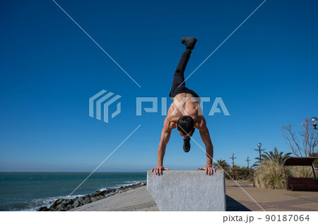Shirtless man doing a handstand on a parapet by the sea.  90187064