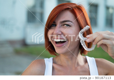 Red-haired Caucasian woman holding transparent mouthguards for bite correction outdoors. A girl with a beautiful snow-white smile uses silicone braces Red-haired Caucasian woman holding transparent mouthguards for bite correction outdoors. A girl with a beautiful snow-white smile uses silicone braces 90187113