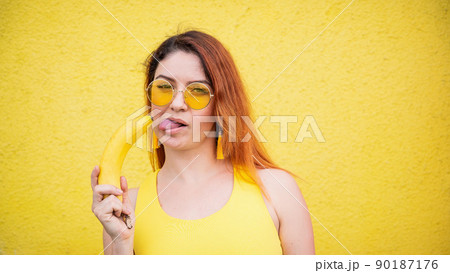 A beautiful red-haired woman in a yellow dress and sunglasses shows her tongue and and licks a banana. Portrait of a fantasizing girl with ripe fruit. 90187176