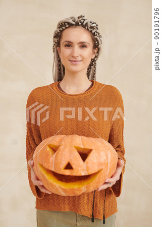 Portrait of content attractive young woman in autumn sweater holding big pumpkin with carved face against isolated background 90191796