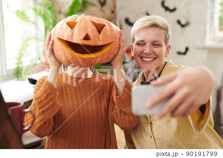 Smiling young woman with blond hair taking funny selfie with friend covering face with carved pumpkin at Halloween party 90191799