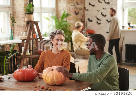 Cheerful young interracial friends in sweaters sitting at table in lift studio and carving pumpkins for Halloween party Cheerful young interracial friends in sweaters sitting at table in lift studio and carving pumpkins for Halloween party 90191820
