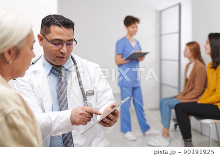 Middle-Eastern doctor showing test results to woman while his colleague calling for next patient in queue 90191925