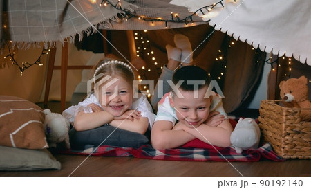 A little boy and girl lie in a tent of blankets and pillows in the bedroom and look at the camera. Children A little boy and girl lie in a tent of blankets and pillows in the bedroom and look at the camera. Children 90192140
