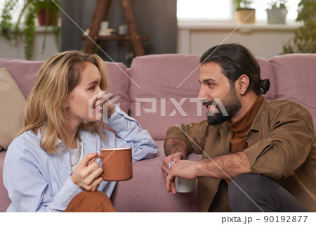Medium close-up portrait of joyful couple sitting on floor against sofa in living room drinking tea and discussing something 90192877