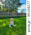 A small, happy, smiling baby in a pink hat, panama, sneakers sits on the grass in a village, a country house. Blue eyes. Against the backdrop of a fence 90198277
