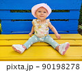 A small, smiling, happy, blue-eyed girl, a baby sits on a yellow-blue bench, the color of the Ukrainian flag. In the park. Pink hat, panama, pants, sneakers. Summer, spring, day 90198278