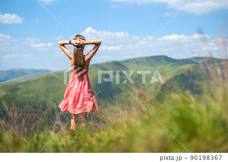 Young woman in red dress standing on grassy field on a windy day in summer mountains enjoying view of nature. 90198367