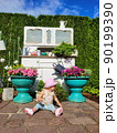 A little girl, a baby in a pink hat, panama against the backdrop of a cabinet installation with pots and flowers, bushes, grass in the garden, sits on the stoves. Looking away 90199390