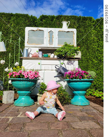 A little girl, a baby in a pink hat, panama against the backdrop of a cabinet installation with pots and flowers, bushes, grass in the garden, sits on the stoves. Looking away 90199390