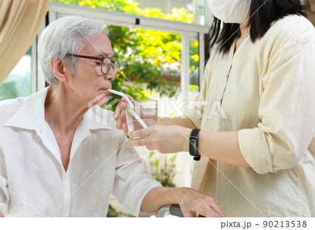 Asian caregiver take care of senior woman give drinking water from a glass,feeding water using a straw to prevent choking at nursing home,thirsty elderly patient sucking water through straw for safety 90213538