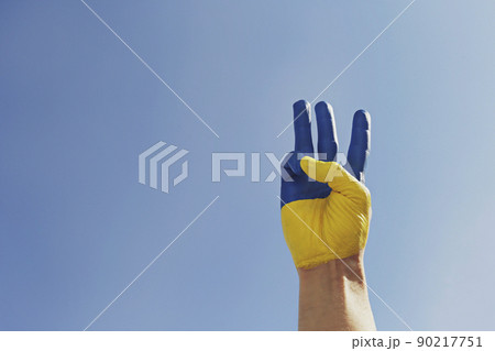 Man's hand lit by sunshine painted in colors of Ukrainian national flag showing a traditional trident hand gesture as a symbol of support and solidarity with Ukraine. Copy space, close up, background. Man's hand lit by sunshine painted in colors of Ukrainian national flag showing a traditional trident hand gesture as a symbol of support and solidarity with Ukraine. Copy space, close up, background. 90217751