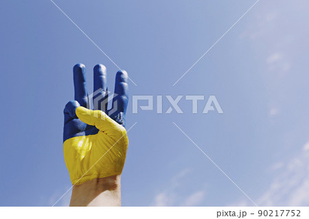 Man's hand lit by sunshine painted in colors of Ukrainian national flag showing a traditional trident hand gesture as a symbol of support and solidarity with Ukraine. Copy space, close up, background. Man's hand lit by sunshine painted in colors of Ukrainian national flag showing a traditional trident hand gesture as a symbol of support and solidarity with Ukraine. Copy space, close up, background. 90217752